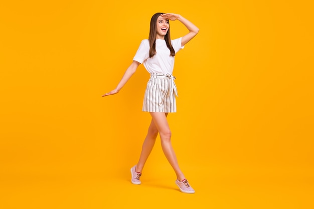 Foto de perfil de cuerpo completo de una atractiva dama divertida caminando por la calle disfrutar de un día soleado, mirar lejos, usar camiseta blanca casual, pantalones cortos a rayas, zapatos aislados, pared de color amarillo brillante