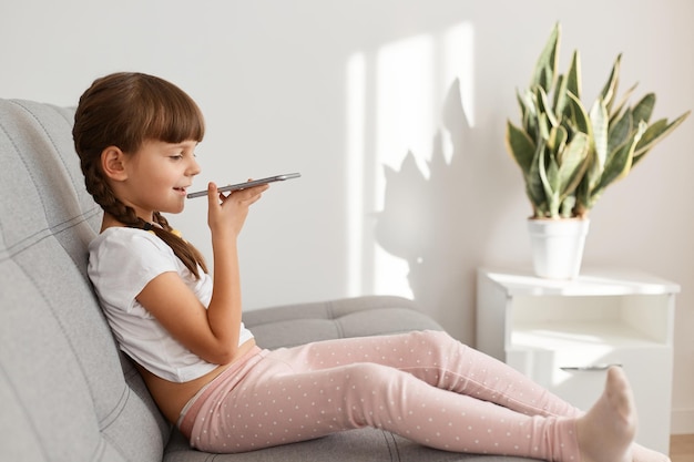 Foto de perfil en interiores de adorable niña con coleta sentada en el sofá en la sala de estar luminosa con el teléfono inteligente en las manos, grabando mensajes de voz para el mejor amigo o los padres.