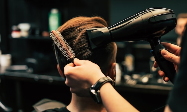 Foto de perfil de un joven peluquero recortando el cabello de su cliente con una afeitadora eléctrica y un peine en una barbería.