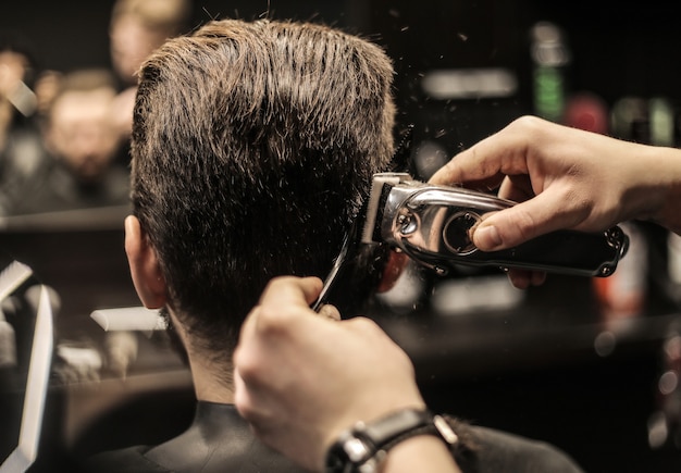 Foto foto de perfil de un joven peluquero recortando el cabello de su cliente con una afeitadora eléctrica y un peine en una barbería.