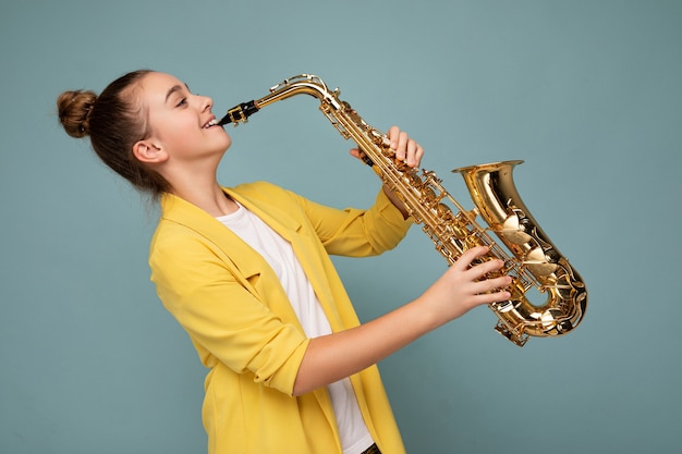 Foto de perfil lateral de una adolescente morena sonriente bastante positiva con chaqueta amarilla de moda que se encuentran aisladas sobre la pared de fondo azul tocando el saxofón mirando hacia el lado