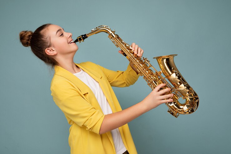 Foto foto de perfil lateral de una adolescente morena sonriente bastante positiva con chaqueta amarilla de moda que se encuentran aisladas sobre la pared de fondo azul tocando el saxofón mirando hacia el lado
