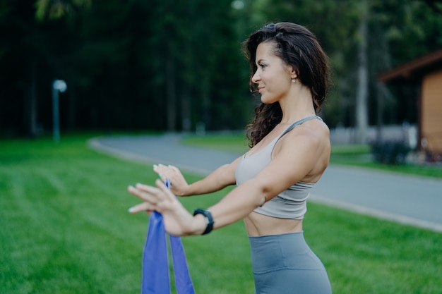 Foto de perfil de mujer delgada deportiva tiene banda de resistencia, tiene entrenamiento físico, vestida con ropa deportiva, posa al aire libre contra el fondo de la naturaleza. Concepto de personas, estilo de vida, stetching y ocio