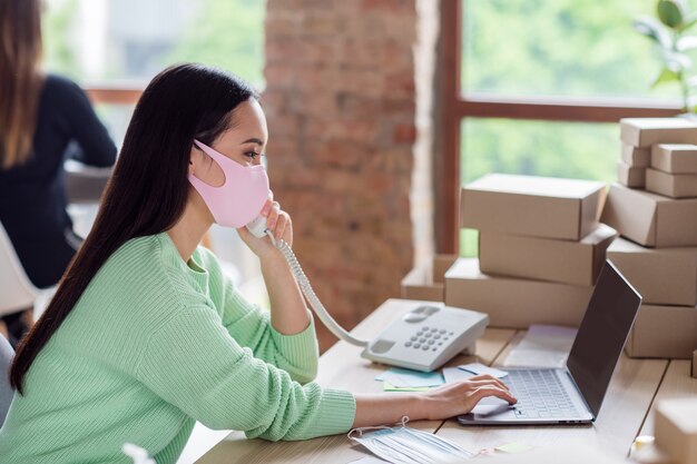 Foto de perfil de la mujer de negocios asiática gerente que organiza los paquetes de máscaras médicas para la gripe facial cajas de entrega que hablan los detalles del pedido del cliente de la línea fija anotando información en el cuaderno de la oficina en casa en el interior