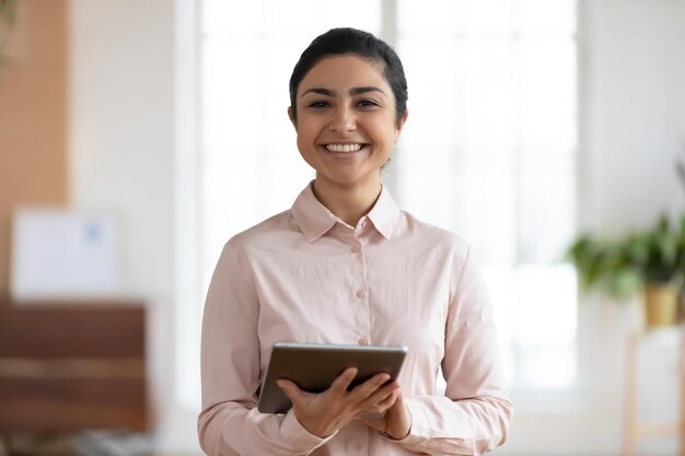 Foto de perfil de una sonriente mujer de negocios india milenaria que usa un moderno dispositivo de tableta en la oficina