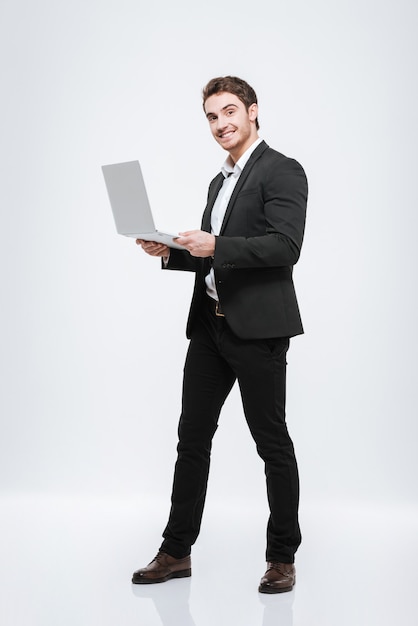 Foto de pie alegre joven empresario caucásico. Aislado sobre pared blanca. sosteniendo la computadora portátil en las manos.