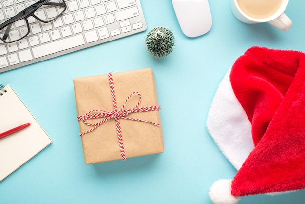 Foto plana de vista superior de caja de regalo con lazo de cuerda sombrero de santa claus teclado computadora mouse gafas bloc de notas pluma abeto juguete y taza de bebida caliente sobre fondo azul pastel aislado