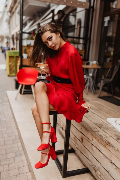 Foto en pleno crecimiento de la hermosa morena Chica en vestido escarlata romántica y linda sonrisa en la lente mientras está sentada en el café Mujer disfrutando de un buen día mientras bebe vino