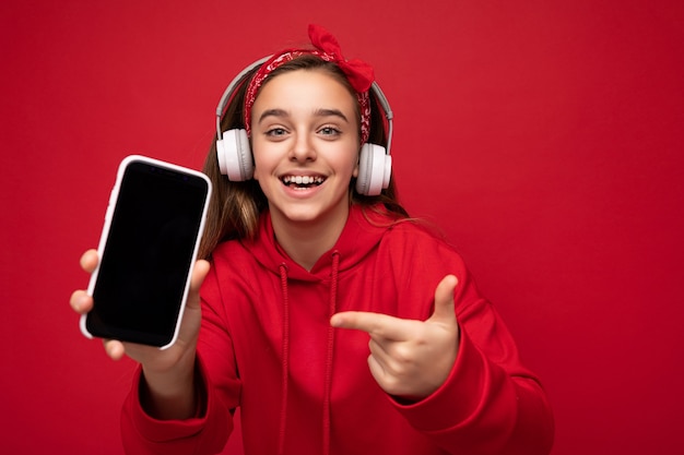 Foto de positivo sonriente linda chica morena vestida con capucha roja aislado sobre fondo rojo sosteniendo