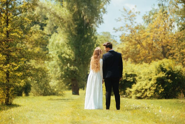 La foto posterior de una novia y un novio tomados de la mano y mirándose al aire libre en el parque o bosque.