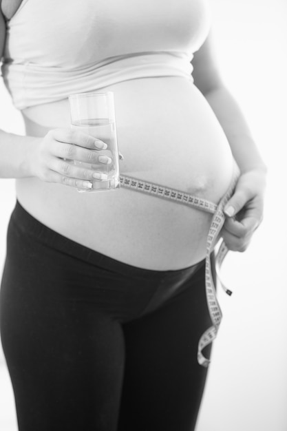 Foto de primer plano en blanco y negro de la mujer embarazada sosteniendo un vaso de agua y midiendo la barriga grande