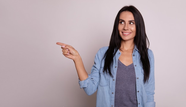 Foto de primer plano de una chica encantadora con cabello largo y oscuro, que está sonriendo y posando con los brazos cruzados, apuntando con su mano izquierda a la esquina superior izquierda