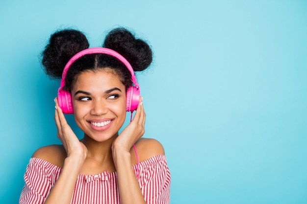 Foto de primer plano de una dama de piel oscura interesada divertida escuchar música auriculares modernos mirar espacio vacío lateral usar camisa a rayas blanca roja de moda fuera de los hombros pared de color azul aislado