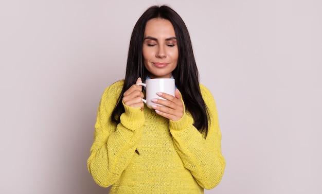 Foto de primer plano de la encantadora morena, que está posando con un suéter amarillo, mirando a un lado y sonriendo mientras sostiene la taza de té blanco