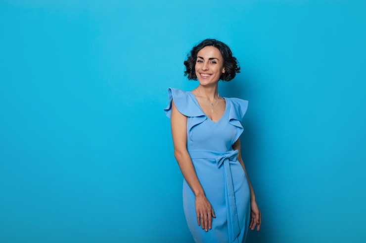 Foto foto de primer plano de una encantadora mujer sana con un vestido azul, que mira a la cámara y sonríe, mientras está de pie y posa aislada en el fondo azul