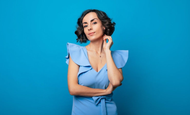 Foto foto de primer plano de una encantadora mujer sana con un vestido azul, que mira a la cámara y sonríe, mientras está de pie y posa aislada en el fondo azul