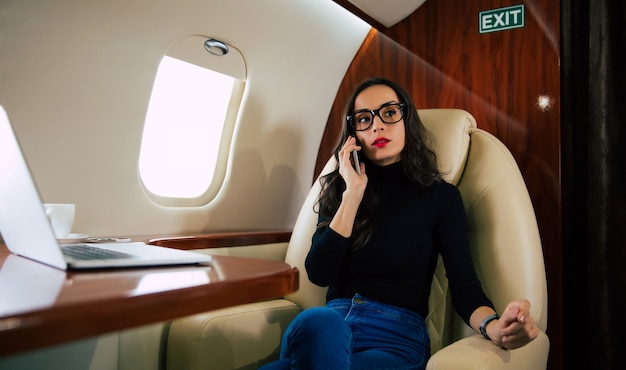 Foto de primer plano de una hermosa mujer con un traje casual, que está hablando por teléfono y bebiendo café solo durante su vuelo en jet privado.