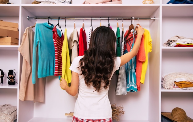 Foto de primer plano de una hermosa niña de cabello castaño largo, que está de espaldas a la cámara, tocando dos prendas de su gran armario.