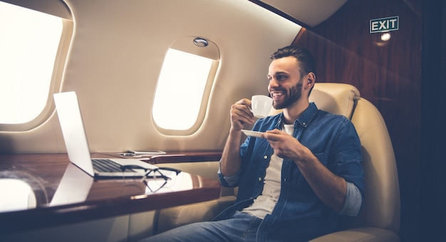 Foto de primer plano de un hombre guapo con un atuendo informal, que está sonriendo, mientras escribe algo en su computadora portátil y sostiene un teléfono inteligente en su mano izquierda, volando en primera clase de avión.