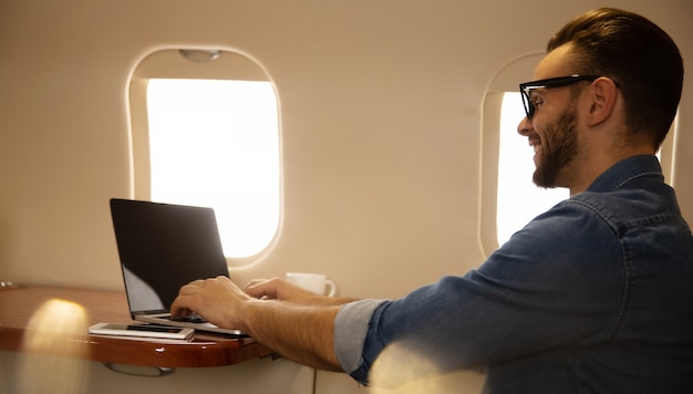 Foto de primer plano de un hombre guapo con un atuendo informal, que está sonriendo, mientras escribe algo en su computadora portátil y sostiene un teléfono inteligente en su mano izquierda, volando en primera clase de avión.