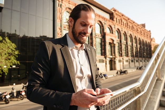 Foto en primer plano de un hombre guapo europeo de 30 años en traje formal con teléfono inteligente, mientras está de pie frente al edificio de oficinas o centro de negocios