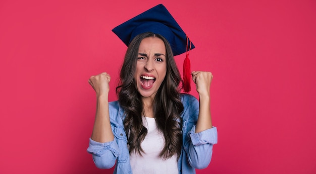 Foto de primer plano de una linda estudiante con una blusa y un sombrero académico cuadrado, que está muy contenta y grita de felicidad por su éxito