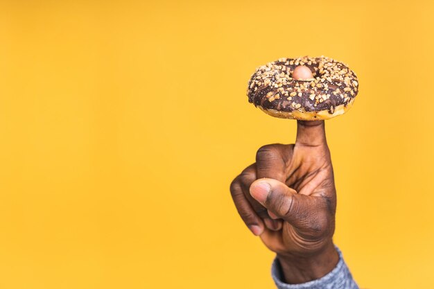 Foto de primer plano de la mano del hombre negro afroamericano hambriento sosteniendo donut aislado sobre fondo amarillo. Comida chatarra. Concepto de dieta.