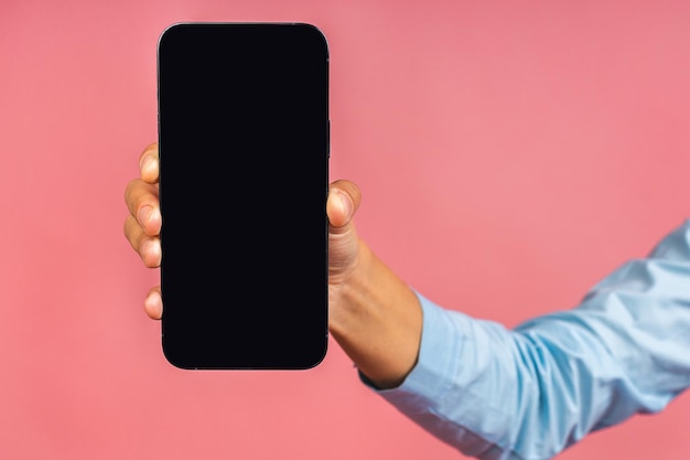 Foto de primer plano de la mano de una mujer afroamericana sosteniendo un teléfono móvil con pantalla en blanco aislado sobre fondo rosa