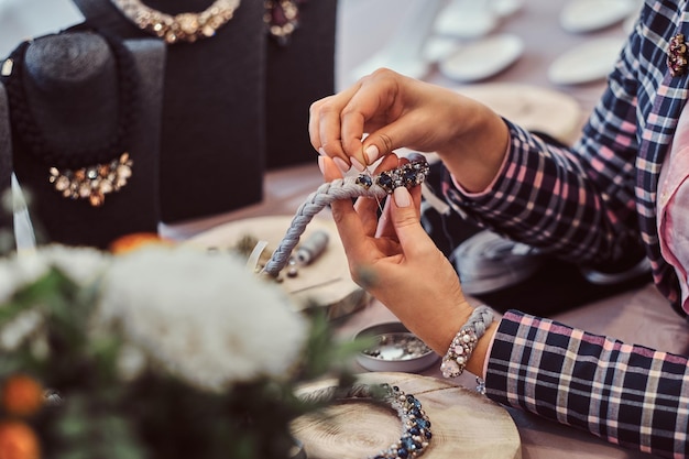 Foto de primer plano de las manos de una mujer que hace collares hechos a mano, trabajando con agujas e hilo en un taller de joyería.
