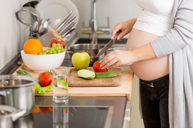 Foto en primer plano de la mujer embarazada cortando verduras sobre tabla de madera