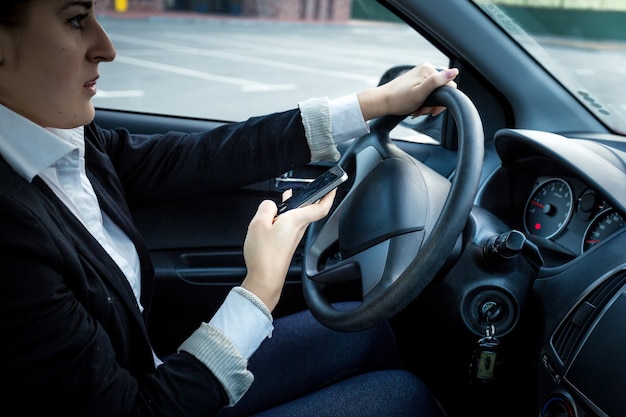 Foto en primer plano de una mujer joven conduciendo un coche y escribiendo un mensaje