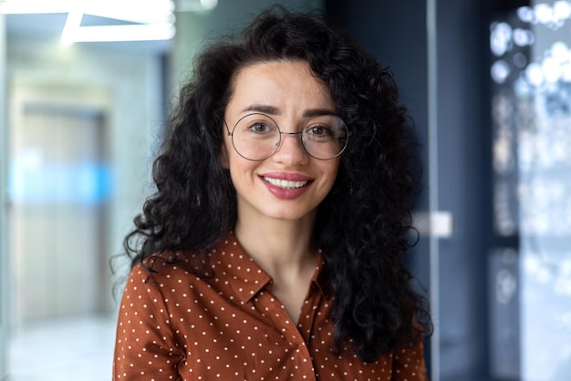 Foto de primer plano de una mujer de negocios con gafas y cabello rizado sonriendo y mirando el retrato de la cámara