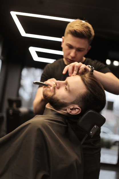 Foto de primer plano de un peluquero profesional, que arregla la barba de su joven cliente, que se mira en el espejo durante el procedimiento.