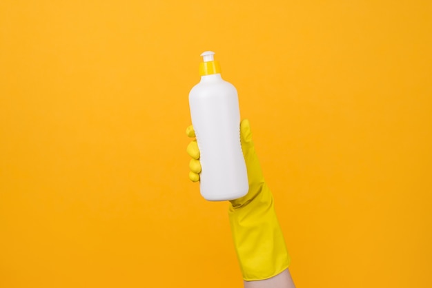 Foto en primer plano recortada de la mano en el guante amarillo sosteniendo una botella de detergente sobre fondo amarillo