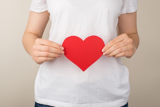 Foto de primer plano recortada de una mujer con camiseta blanca y jeans sosteniendo un corazón de papel rojo sobre un fondo gris aislado