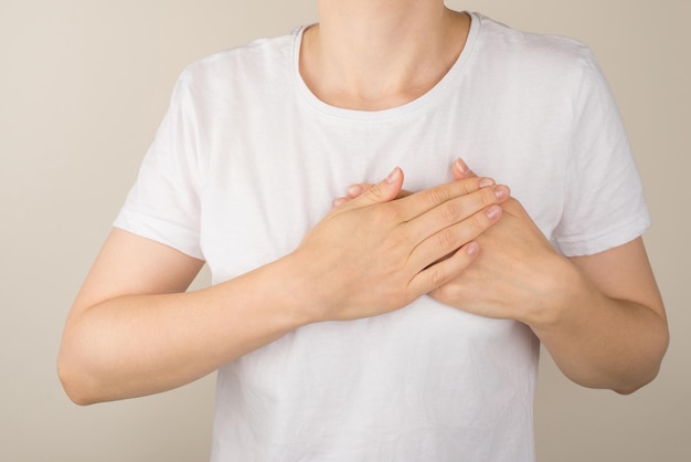 Foto de primer plano recortada de una mujer con camiseta blanca sosteniendo sus manos en el corazón sobre un fondo gris aislado