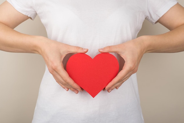 Foto de primer plano recortada de una mujer joven con camiseta blanca sosteniendo un corazón de papel rojo cerca de la parte superior del abdomen sobre un fondo gris aislado