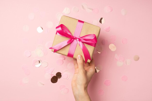 Foto en primera persona de la mano de una chica sosteniendo una elegante caja de regalo de papel artesanal con un lazo de cinta de raso rosa sobre confeti brillante en un fondo rosa pastel aislado