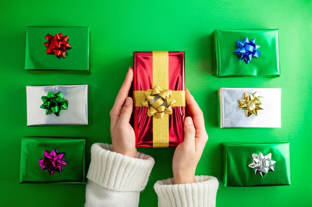 Foto en primera persona de la mano sosteniendo una caja de regalos roja con cinta de satén amarillo sobre mini regalos de colores en un fondo verde aislado con copyspacexA