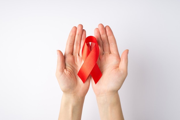 Foto en primera persona de las manos de una mujer joven sosteniendo una cinta de seda roja en las palmas, símbolo de concienciación sobre el sida en un fondo blanco aislado