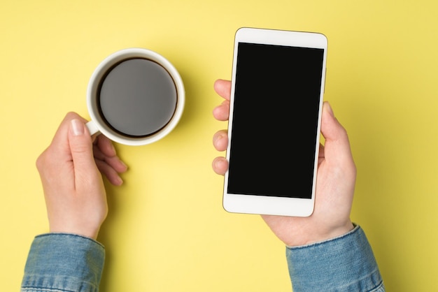 Foto en primera persona de las manos de una mujer sosteniendo una taza de café y un teléfono inteligente en un fondo amarillo aislado con espacio de copia