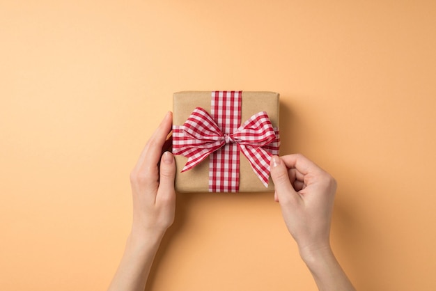 Foto en primera persona de la vista superior de las decoraciones del día de san valentín manos de una mujer joven desatando un lazo de cinta a cuadros en una caja de regalo de papel artesanal sobre un fondo beige aislado