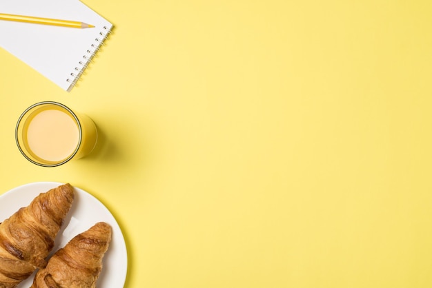 Foto foto de primera vista de un vaso de plato de jugo con cruasanes recién hechos y lápiz amarillo en un cuaderno pequeño sobre fondo amarillo pastel aislado con espacio de copia