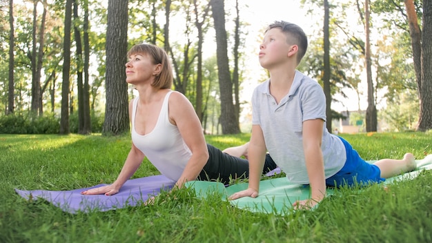 Foto de una profesora de yoga de mediana edad o un gurú que enseña a un adolescente que hace yoga. Mujer con niño meditando y estirando sobre el césped en el parque