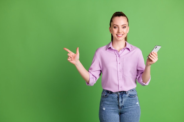 La foto del promotor de la niña positiva usa el copyspace del dedo índice del punto del teléfono inteligente indica la promoción de las redes sociales usa camisa lila violeta jeans de mezclilla aislados sobre fondo de color verde