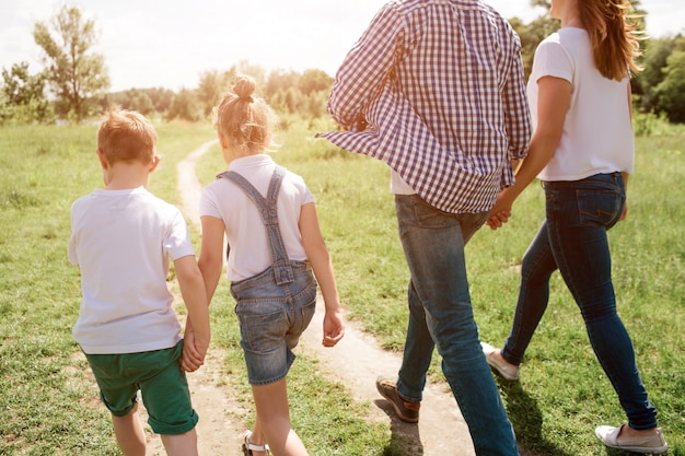 Una foto que fue hecha desde atrás. Familia feliz es caminar juntos a través del Prado. El muchacho está sosteniendo la mano de su hermana mientras que el hombre está sosteniendo la mano de su esposa. Su andar es confiado.