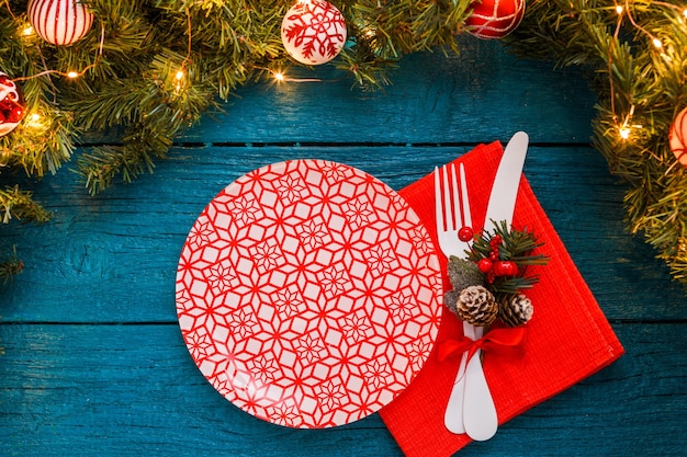 Foto de ramas de pino, platos con patrón rojo, galletas de predicción, tenedor, cuchillo, decoración navideña sobre fondo de madera azul