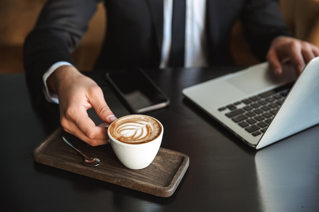 Foto recortada de un apuesto joven empresario sentado en la cafetería con ordenador portátil tomando café.