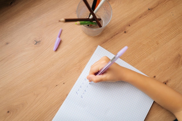 Foto recortada de close-up de garotinha primária irreconhecível fazendo lição de casa, escrevendo no caderno com caneta, sentado à mesa. Vista de alto ângulo da escola elementar menina aprendendo em casa.