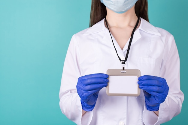 Foto recortada de close-up de médica em luvas de uniforme de perigo de jaleco branco segurando um crachá maok em branco isolado sobre fundo azul